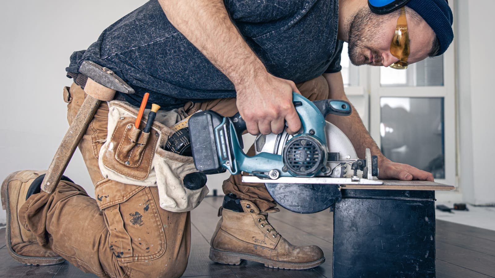 Tradesperson in safety goggles and a tool belt kneeling to cut a board with a circular saw