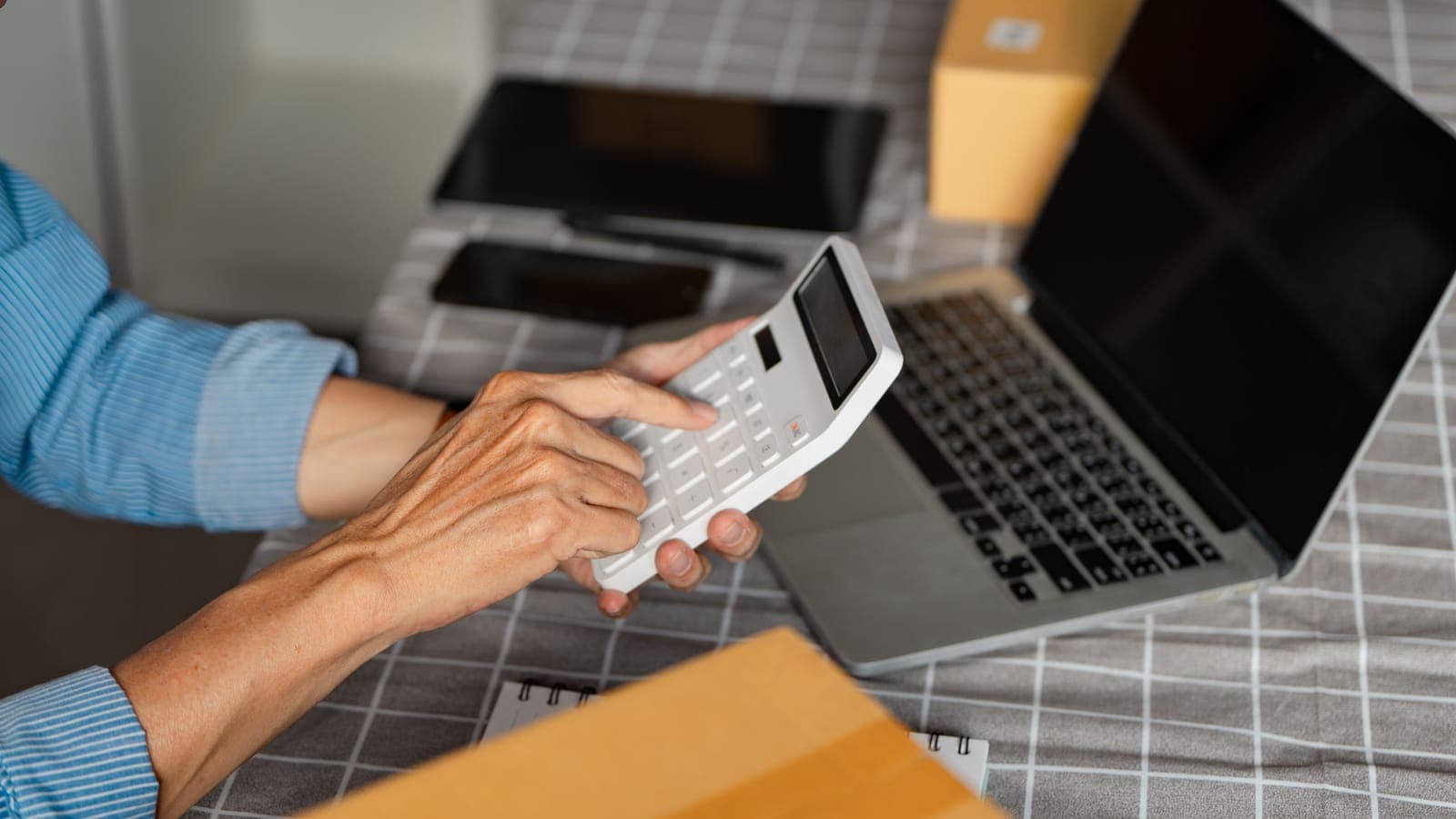Hands using a white calculator beside a laptop and parcels, working out business finances
