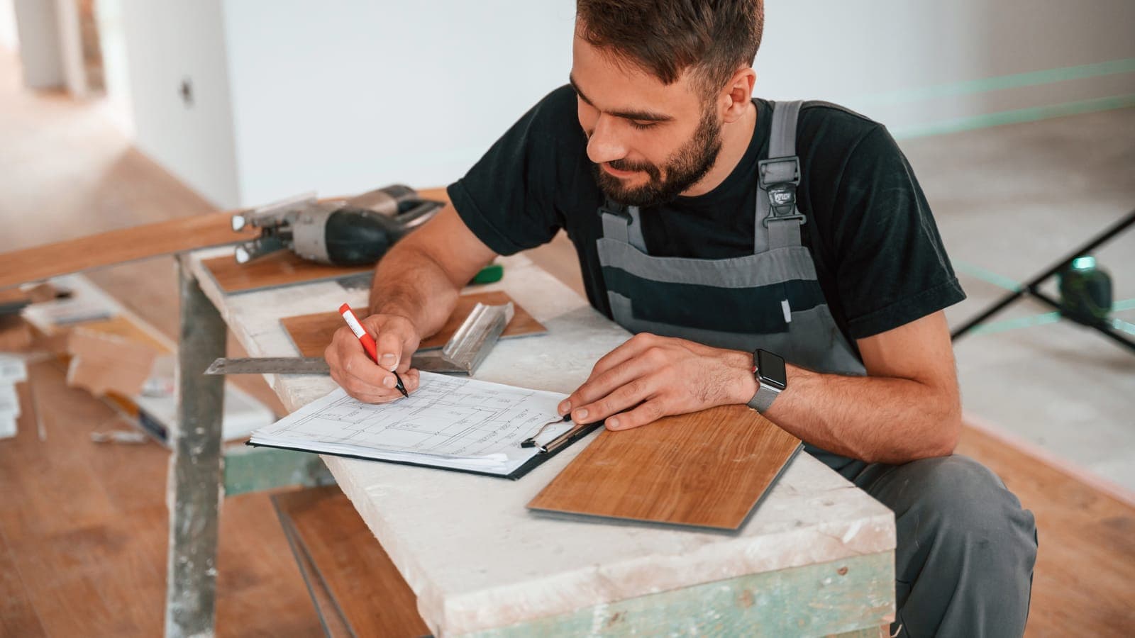 Tradesperson in overalls writing a job quote on paperwork at a workbench with timber samples