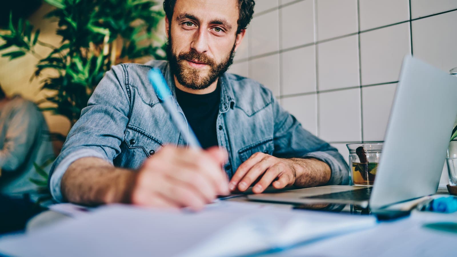 Bearded man in a denim shirt writing notes beside a laptop at a cafe table