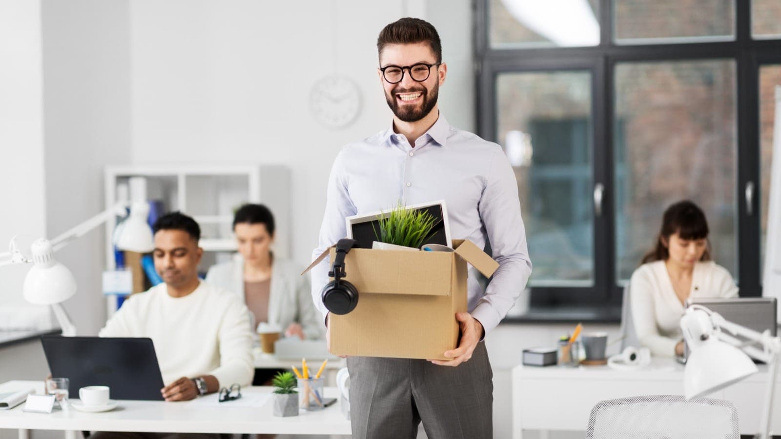 Man in an open-plan office holding a box of desk items on his last day