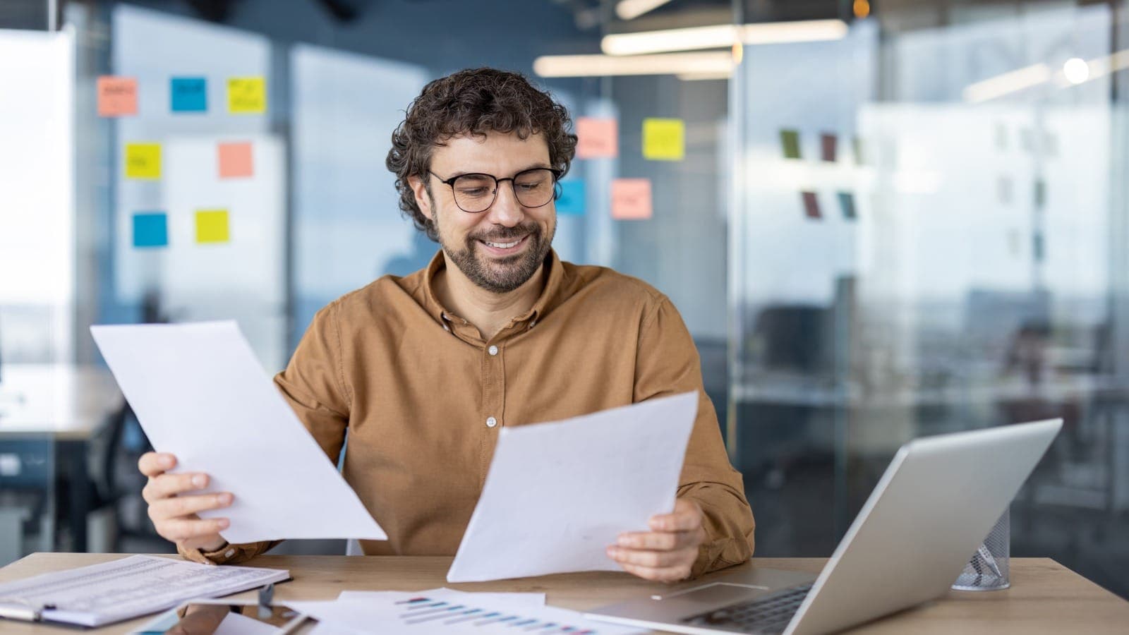 Man at a desk comparing two pages of paperwork beside a laptop, sticky notes behind