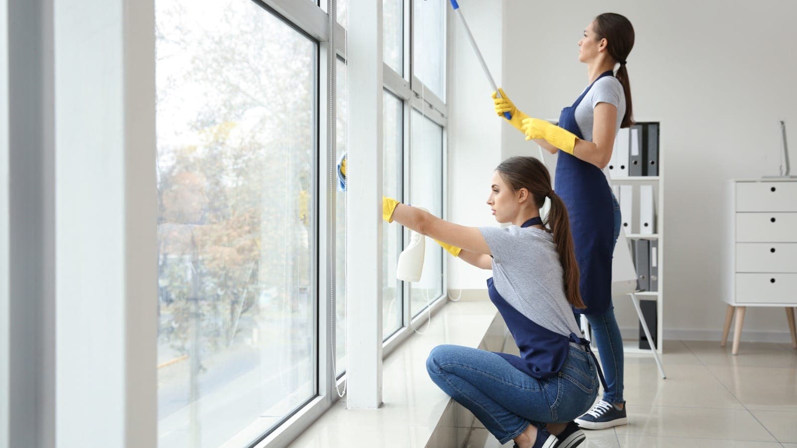 Two window cleaners using a squeegee and spray on a large interior window
