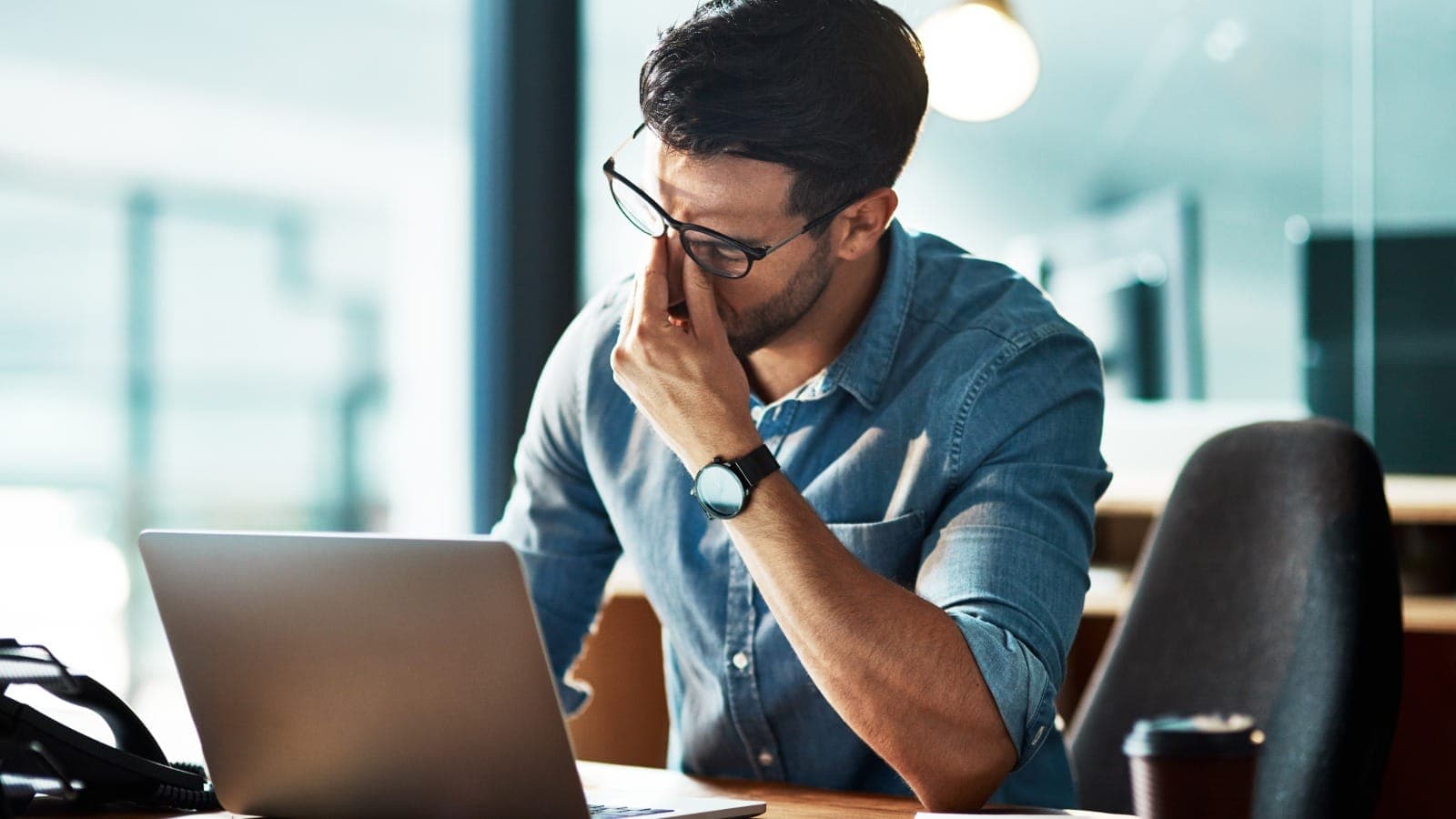 Man with glasses leaning his head on his hand at a laptop, eyes closed in frustration