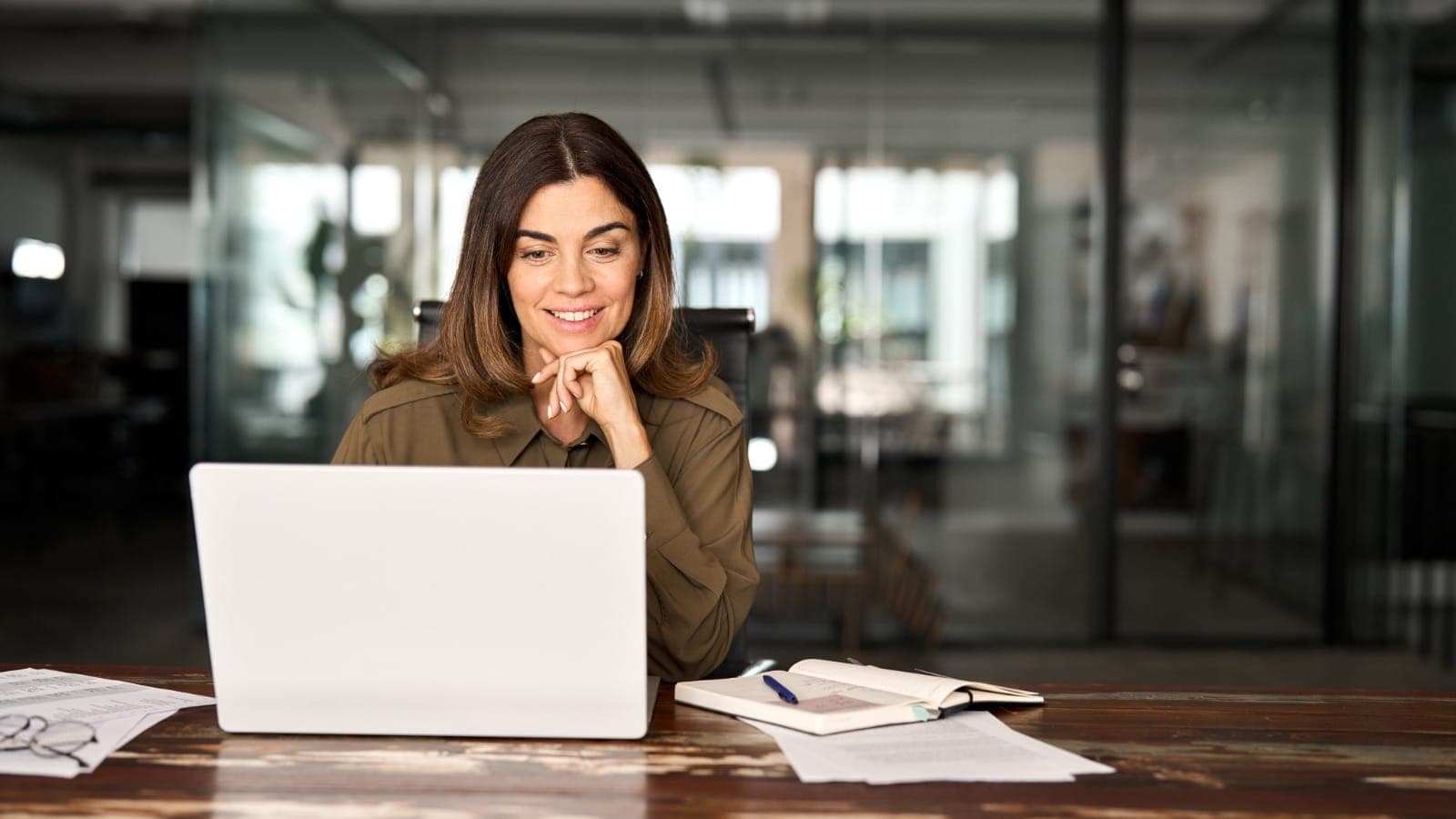 Smiling woman at a laptop with a notebook and papers in a modern office