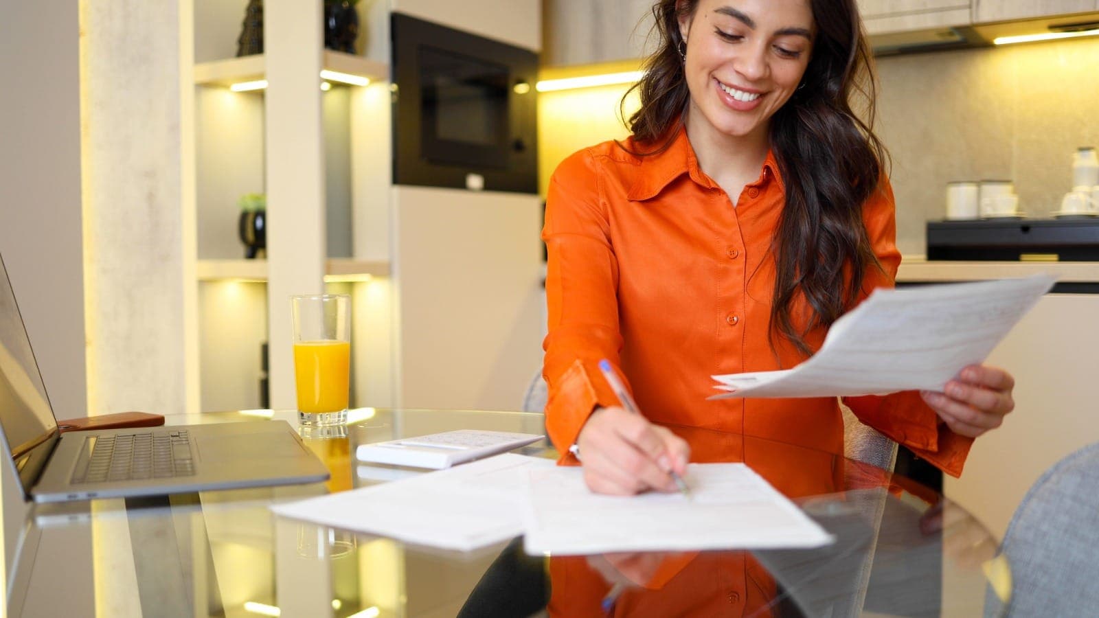 Smiling woman writing on paperwork at home with a laptop, calculator and notebook